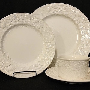 May include: A set of white ceramic dinnerware with a grapevine design. The set includes a dinner plate, a salad plate, and a teacup and saucer.