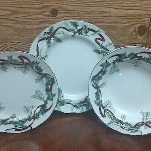 May include: Three white ceramic plates with a festive holiday design. Each plate features a green holly and red ribbon border. The plates are arranged on a brown surface against a wooden background.