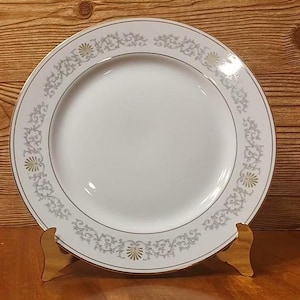 May include: White ceramic plate with a decorative border featuring a floral and scroll design in grey and gold. The plate has a gold trim and is displayed on a wooden stand. A classic piece for serving.