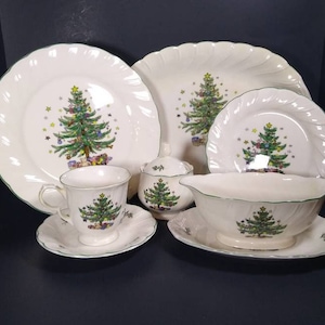 May include: A cream-colored Christmas dinnerware set featuring a festive Christmas tree design. The set includes plates, a serving platter, a gravy boat, a sugar bowl, and a teacup with saucer. Each piece has a green trim and a scalloped edge.