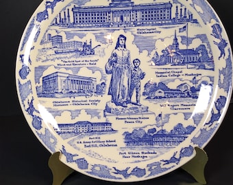 Könnte beinhalten: Ein runder, weiß-blauer Zierteller mit Illustrationen von Wahrzeichen von Oklahoma. Der Teller zeigt Bilder des State Capitol, der Memorial Chapel und der Pioneer Woman Statue. Der Teller wird von einem goldfarbenen Ständer getragen.