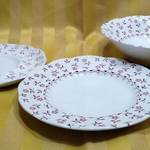 May include: Three white ceramic plates with a floral pattern in shades of pink and purple. The plates are all different sizes, with the largest plate in the center and the smallest plate on the left.
