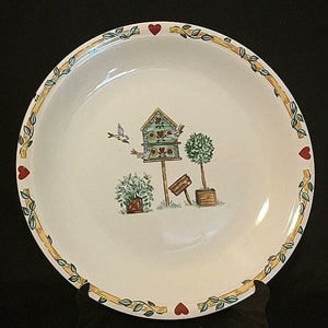 May include: A white ceramic pie plate with a yellow and green border featuring a birdhouse, birds, and plants. The border is decorated with red hearts.