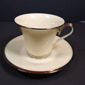 Könnte beinhalten: Eine weiße Porzellan-Teetasse und -Untertasse mit goldener Verzierung. Die Tasse hat eine abgerundete Form und einen Henkel. Die Untertasse ist rund und hat einen leicht erhöhten Rand.