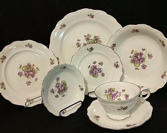 May include: A set of white porcelain dinnerware with a delicate floral pattern featuring purple violets. The set includes a large dinner plate, a salad plate, a soup bowl, a teacup, and a saucer.