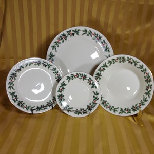 May include: Set of four white ceramic plates with a green holly and red berry border. The plates are different sizes, with the largest plate in the back and the smallest plate in the front.