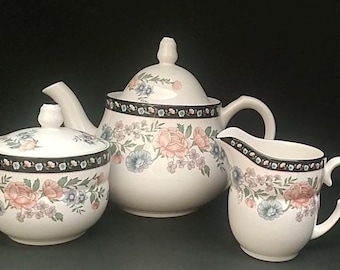 May include: A white ceramic tea set consisting of a teapot with a lid, a sugar bowl with a lid, and a creamer. Each piece features a black border with a floral pattern of pink roses and blue flowers around the centre and upper rim.