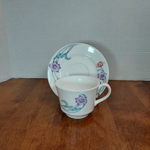 May include: A white teacup and saucer set with a floral design. The cup and saucer feature a green, purple, and pink floral pattern.