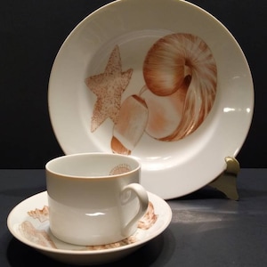 May include: A white ceramic dish set featuring a seashell design. The set includes a large plate, a teacup, and a saucer. The seashells are depicted in shades of brown and tan, creating a coastal theme.