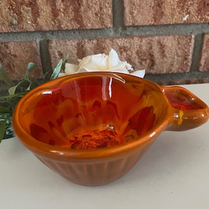 May include: A small, orange ceramic bowl with a handle. The bowl has a glazed finish and a textured exterior.