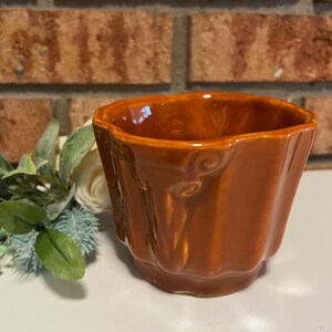 May include: A small, brown ceramic planter with a textured, tree bark-like design. The planter has a scalloped rim and is perfect for small plants.