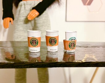 May include: Three miniature Starbucks coffee cups with brown sleeves, each featuring the Starbucks logo. The cups are white with a gold rim. The cups are on a dark, reflective surface.