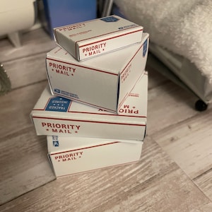 May include: A stack of five white cardboard boxes with blue and red trim. Each box has the text "PRIORITY MAIL" printed on it. The boxes are stacked on a wooden surface.