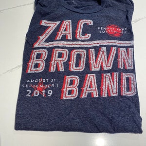 May include: A navy blue t-shirt with white and red lettering that reads "Zac Brown Band" with the dates "August 31" and "September 1, 2019" and the text "Fenway Park Boston, MA".