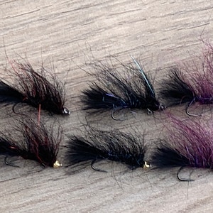 May include: Six handmade fishing flies. Each fly features a black body with a fluffy, feather-like texture, a curved hook, and a small, metallic bead. Some flies have purple or red accents.