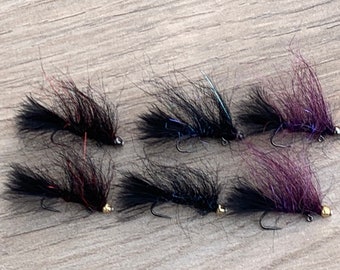 May include: Six handmade fishing flies. Each fly features a black body with a fluffy, feather-like texture, a curved hook, and a small, metallic bead. Some flies have purple or red accents.