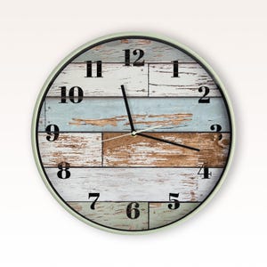 May include: A round wall clock with a light green frame and a face featuring horizontal planks of distressed wood in shades of white, light blue, and brown. The clock has black Arabic numerals from 1 to 12 and thin black hour and minute hands.