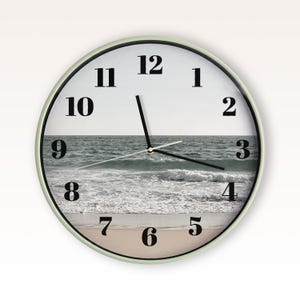 May include: A round wall clock with a light green frame and a white face featuring a photographic print of an ocean shoreline. The clock has black serif numerals from one to twelve and three thin black hands indicating the current time.