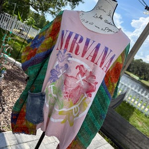 May include: A light pink t-shirt featuring the word NIRVANA in a rainbow gradient font above a white graphic of a smiley face overlaid with floral patterns. The shirt has long, multicolored patchwork sleeves with visible stitching and a denim pocket.