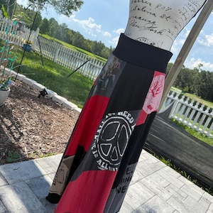 May include: A high-waisted skirt featuring a black waistband and a multi-panel design with red and black sections. The fabric displays various graphic prints, including a peace sign with the text "ALL YOU NEED IS LOVE" and other patterns.