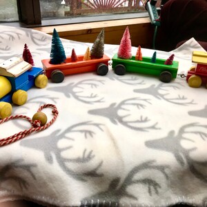 May include: A vintage wooden toy train with a yellow engine, blue caboose, and three green and red boxcars. Each boxcar is filled with small artificial Christmas trees. The train is on a white blanket with a gray deer pattern.
