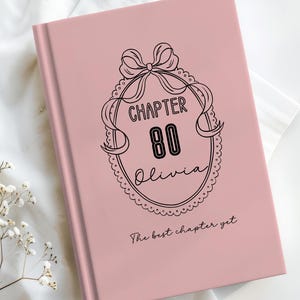 May include: A pink book with the text "CHAPTER 80 Olivia" in a decorative oval frame. The book also has the text "The best chapter yet" at the bottom. The book is on a white surface with flowers.