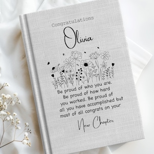 May include: A light gray journal with the name "Olivia" in cursive and the words "Congratulations" and "New Chapter" printed on the cover. A floral design and inspirational text are also featured. The journal is on a white surface.