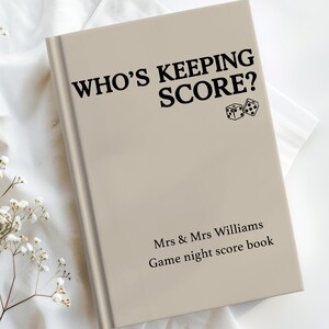 May include: A beige hardcover game night score book with black text that reads WHO'S KEEPING SCORE? along with a small graphic of two dice. Below this, the text reads Mrs & Mrs Williams and Game night score book in black lettering.