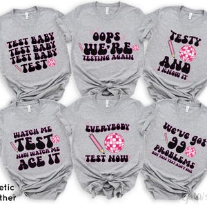 May include: Six gray t-shirts with pink and black text and graphics. The shirts feature phrases like "Test Baby", "Oops We're Testing Again", "Testy and I Know It", "Watch Me Test Now Watch Me Ace It", "Everybody Test Now", and "We've Got 99 Problems But This Test Ain't One".