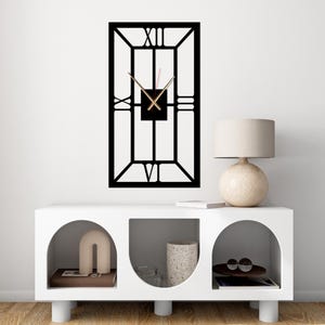 May include: A black rectangular wall clock with Roman numerals. The clock has gold-colored hands and a square center. The clock is mounted on a white wall above a white cabinet with arched openings. A lamp and decorative items are on the cabinet.