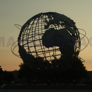 以下が含まれることがあります： 夕焼けのオレンジと黄色の空を背景に、大陸が描かれた大きな金属製の地球儀の彫刻のシルエット。彫刻のフレームワークが見え、前景には木々や公園のような環境があります。