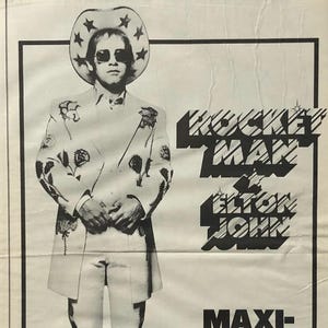 May include: Framed vintage print featuring Elton John in a stage outfit, with the text "Rocket Man" and "Maxi-Single". The image is monochrome, with a white border and a black frame, reminiscent of a music advertisement.