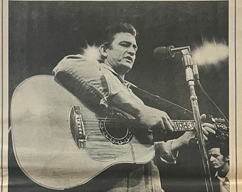 May include: Black and white photo of Johnny Cash performing live at San Quentin prison. The photo shows Cash playing a guitar and singing into a microphone. The text "JOHNNY CASH AT SAN QUENTIN" is printed in bold at the top of the photo.