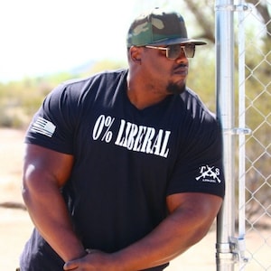 May include: Black t-shirt with the text "0% LIBERAL" in white. The shirt features an American flag on the left sleeve and the "T&S Apparel" logo on the right sleeve. The person is wearing a camouflage baseball cap and sunglasses.