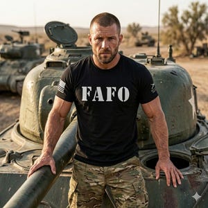 May include: Black t-shirt with the white text "FAFO" and an American flag on the left sleeve. The person is standing in front of a military tank, wearing camouflage pants, in a desert environment.