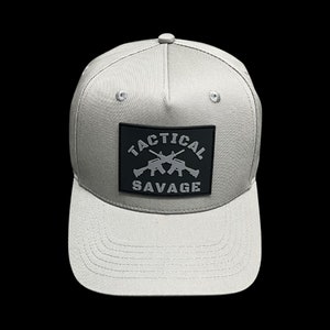 Puede incluir: Gorra de b&eacute;isbol gris claro con visera plana. La gorra presenta un parche cuadrado negro con las palabras "TACTICAL SAVAGE" y dos rifles cruzados en gris. La gorra tiene una corona estructurada y un bot&oacute;n en la parte superior.