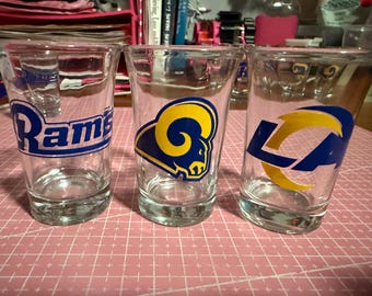 May include: Three clear shot glasses with Los Angeles Rams logos. One glass has the word "Rams" in blue and white. The other two glasses have the team's logo in blue and yellow. The glasses are on a pink grid surface.