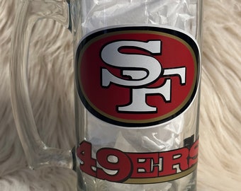May include: Clear glass beer mug with a handle. The mug features the San Francisco 49ers logo in red, gold, and white. The words "49ers" are printed in red on the bottom of the mug.