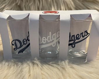 May include: Set of three clear glass shot glasses with a white "Dodgers" logo in a black outline. The glasses are packaged in a white box.