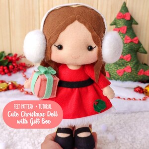 Pode incluir: Boneca de Natal de feltro feita à mão, vestida com um vestido vermelho com cinto preto, acabamento branco e sapatos pretos. A boneca tem cabelo castanho, protetores de ouvido brancos e segura uma caixa de presente rosa com um laço verde. O texto na imagem diz: "FELT PATTERN + TUTORIAL Cute Christmas Doll with Gift Box."