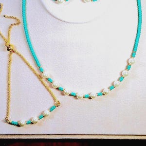 May include: A jewellery set comprising a necklace, bracelet, and earrings. The necklace and bracelet feature white pearls and turquoise beads. The earrings are small pearl studs. All pieces have gold-coloured metal accents.