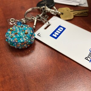 May include: A collection of keys and an ID tag rest on a brown surface. A decorative keychain with blue and gold crystals is attached to the keys. The ID tag has the word "HID" in a blue box.