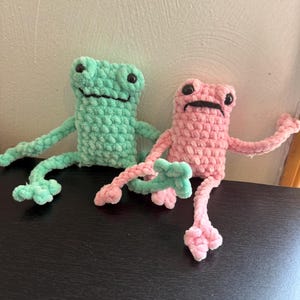 May include: Two handmade crochet frog toys. One frog is light green, the other is pink. Both have black eyes and are sitting on a black surface. The frogs have long arms and legs, and are made of a soft, textured yarn.