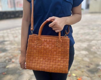 May include: A tan leather handbag featuring a woven square pattern, two rounded top handles, and a long adjustable shoulder strap. The bag has a rectangular shape with visible stitching at the base of the handles and metal hardware attachments.