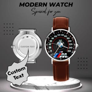 May include: Modern watch with a brown leather strap and a black speedometer-style face featuring the BMW logo. The watch face displays speed markings up to 260 km/h. A second watch with a silver case and space for custom text is also shown.