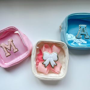 May include: Three small, square, clear plastic cosmetic bags with different colored exteriors. The bags have a zipper closure and a decorative embellishment on the inside. The bag on the left is pink with a gold letter "M" inside. The bag in the center is white with a pink bow inside. The bag on the right is blue with a gold letter "A" inside.