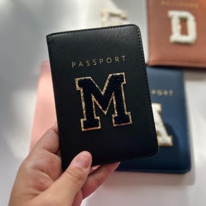 May include: Black passport holder with the word "PASSPORT" and the letter "M" in gold glitter. The letter "M" is surrounded by a black border.
