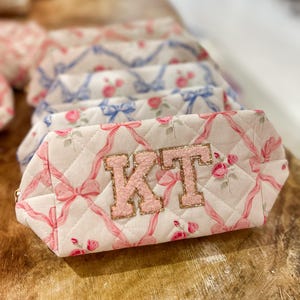 May include: A white quilted makeup bag with pink ribbon and rose print. The bag has a pink and gold glitter monogram "KT" on the front.