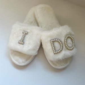 May include: A pair of white fuzzy slippers with the words "I DO" on one slipper and "I" on the other slipper. The letters are decorated with pearl-like embellishments.
