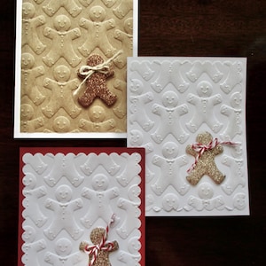 May include: Three rectangular greeting cards featuring embossed gingerbread man patterns. One card is tan with a copper glitter gingerbread man and twine bow. Two cards are white with gold glitter gingerbread men and red and white twine bows.